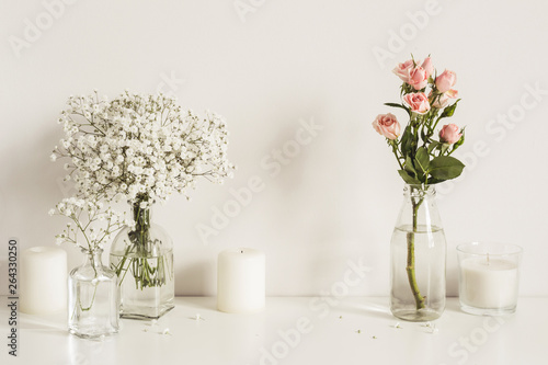 Composition with white and pink flowers in glass bottles and candles on table wall background. Copy space for artwork