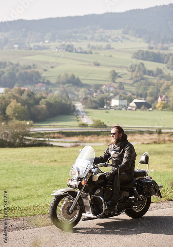 Long-haired bearded cool rider in sunglasses and black leather clothing riding cruiser powerful motorcycle along sunny asphalt road on bright summer day on background of green rural misty landscape.