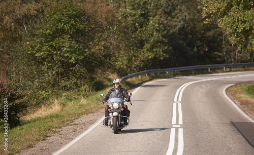 Bearded motorcyclist in helmet  sunglasses and black leather clothing riding bike along empty asphalt road with white marking on bright sunny summer day on background of green dense woody forest.