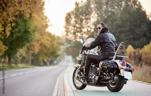 Quadro em tela Back view of handsome bearded motorcyclist in black leather jacket and sunglasses sitting on cruiser motorcycle on blurred background of straight road stretching to horizon and golden autumn trees