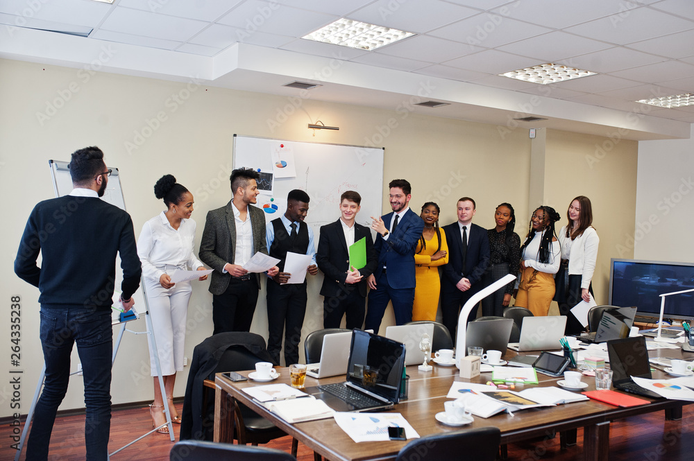 Mixed race business coach presenting report standing near whiteboard pointing on sales statistic shown on diagram and chart teach diverse company members gathered together in conference room.