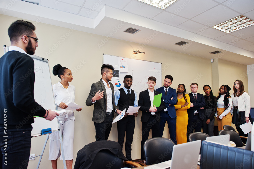 Mixed race business coach presenting report standing near whiteboard pointing on sales statistic shown on diagram and chart teach diverse company members gathered together in conference room.