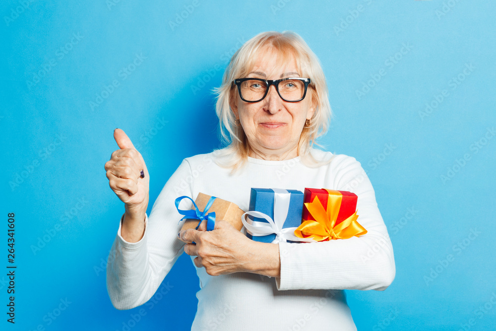 Old woman with a happy facial expression is holding gifts and showing thumbs up gesture against a blue background. Mothers day concept, congratulations, birthday or holiday, approval and gesture Ok. Old woman with a happy facial expression is holding gifts and showing thumbs up gesture against a blue background. Mothers day concept, congratulations, birthday or holiday, approval and gesture Ok.