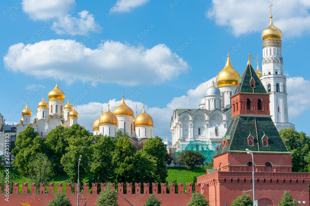 Golden domes of white Orthodox churches behind the walls of the Kremlin against a bright blue sky with white clouds.