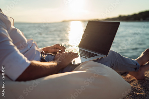 Senior man working on his laptop lying on deck chair on the beach during sunset