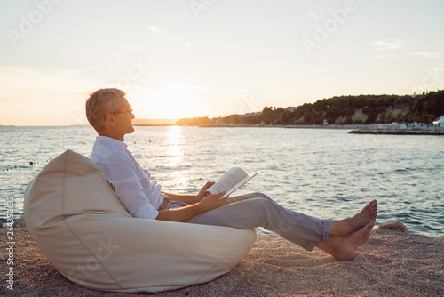 Senior man reading a book lying on deck chair on the beach during sunset