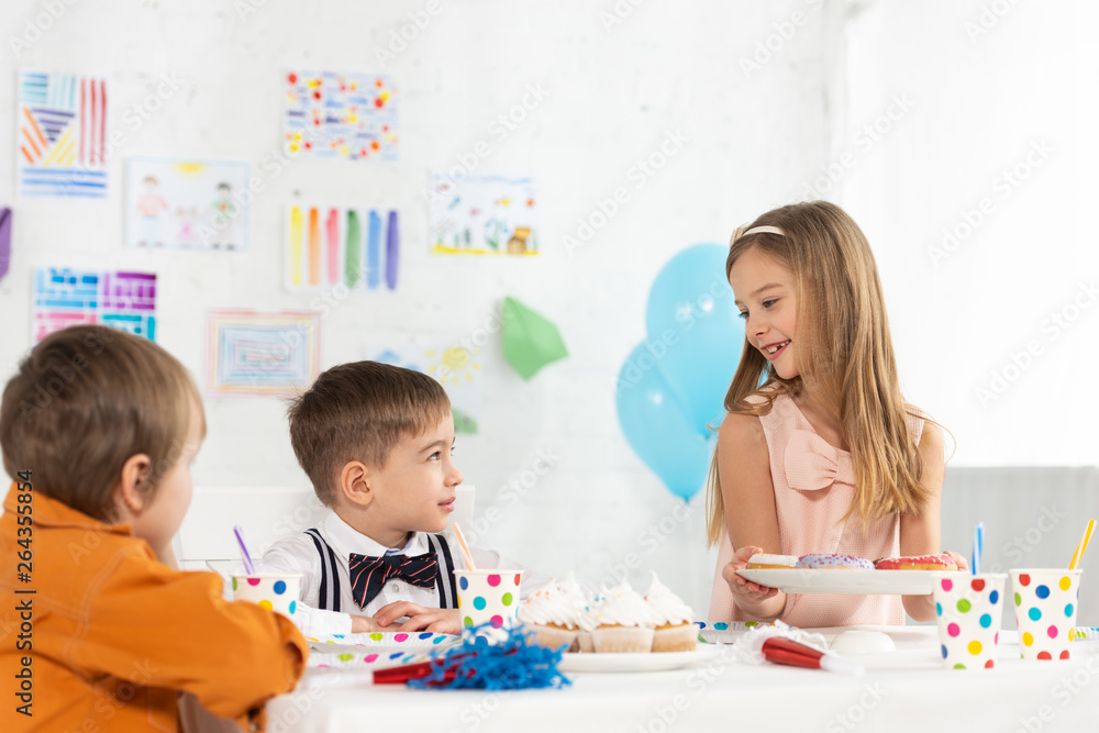 Fototapeta premium smiling kid holding plate with donuts during birthday party with friends at home