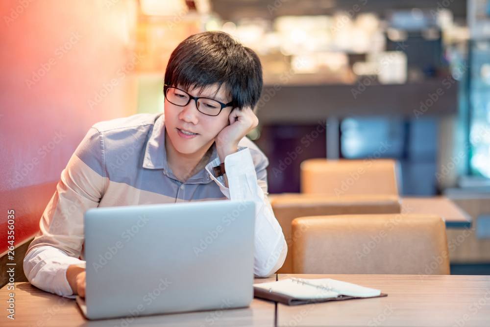 Male Asian businessman wearing glasses working with modern laptop computer on wooden table in the cafe. Work smart in digital age. Internet of things (IOT) concept Male Asian businessman wearing glasses working with modern laptop computer on wooden table in the cafe. Work smart in digital age. Internet of things (IOT) concept