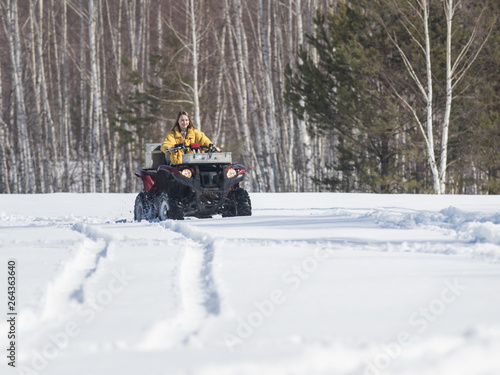 A winter forest. A young smiling woman in bright yellow jacket riding snowmobile