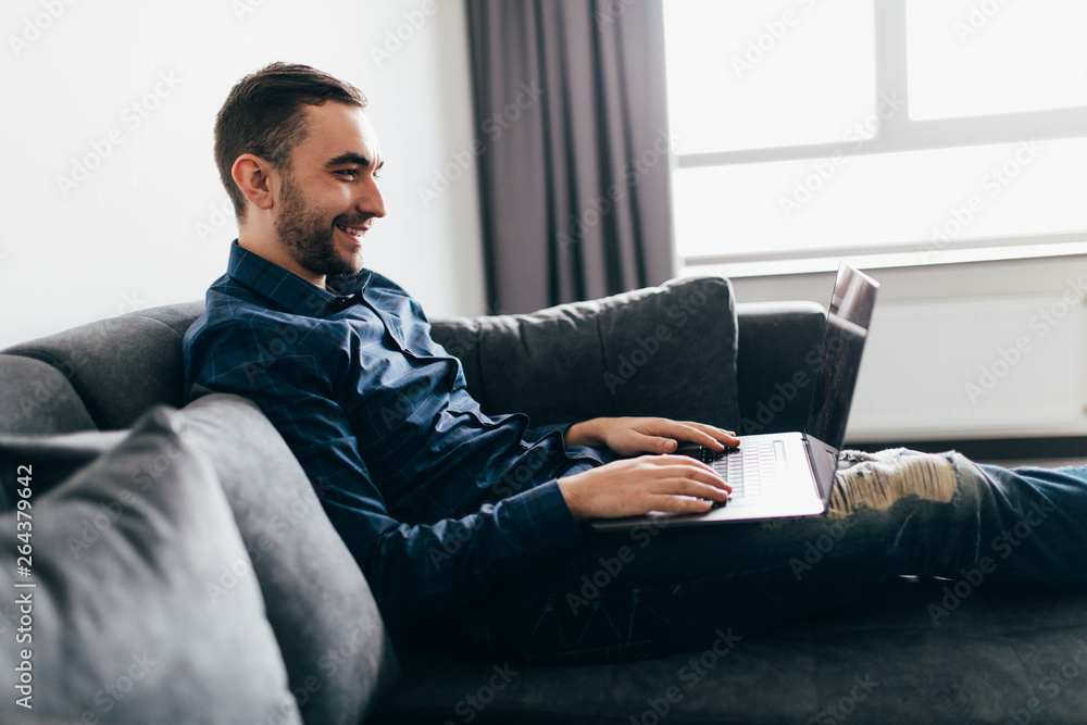 Young attractive smiling man is browsing at his laptop, sitting at home on the cozy beige sofa at home, wearing casual outfit Young attractive smiling man is browsing at his laptop, sitting at home on the cozy beige sofa at home, wearing casual outfit