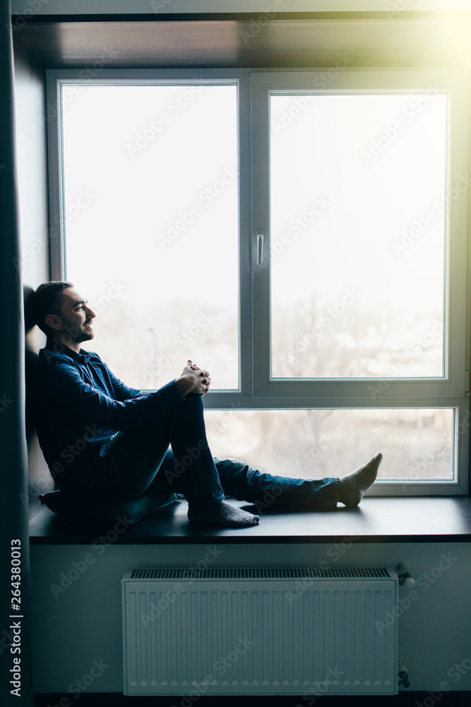Handsome young man sitting on windowsill at home Handsome young man sitting on windowsill at home