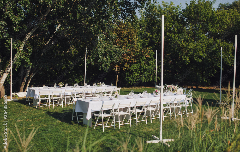 Large white long tables with chairs, decorated with fresh flowers, with plates, glasses and forks, stand in the park with green grass. Wedding decorations and details. Preparing for a wedding party.