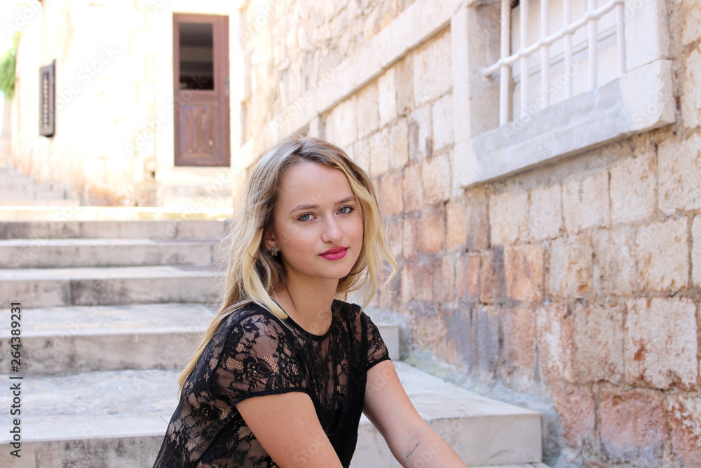 Beautiful romantic young blonde woman with blue eyes in sexy lips sits on steps in a city . She is wearing a black dress. The concept of outdoor relaxing.