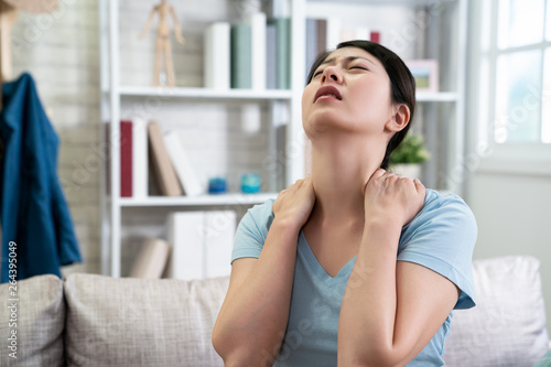 unhappy asian chinese housewife woman has neck pain stretching painful body frowning face. illness japanese wife at home sitting on couch with hurting shoulder. upset female feeling sick indoors.