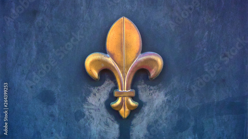 A weather-beaten metal sculpture of the fleur de lis symbol attached to a textured blue wall in horizontal image format.