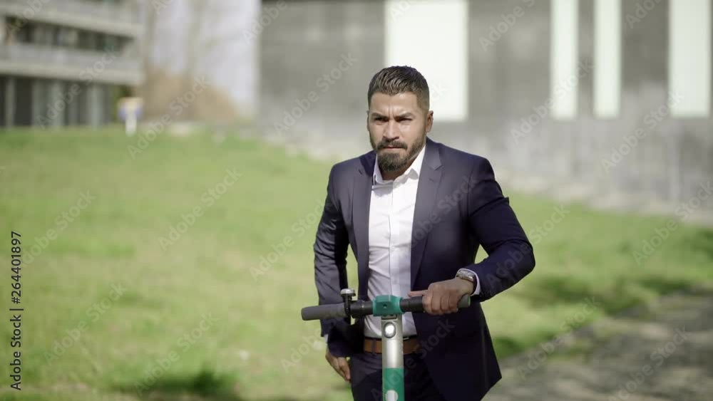Businessman riding scooter and answering phone call. Serious bearded businessman riding scooter on street and using smartphone at sunny day. Transportation concept Businessman riding scooter and answering phone call. Serious bearded businessman riding scooter on street and using smartphone at sunny day. Transportation concept
