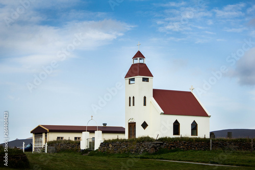 A small catholic church with a tower and brown roof in a small cozy village in Iceland with a nice green garden, green bushes, grass and blue bright sky above. Icelandic minimalistic architecture. © Daniil