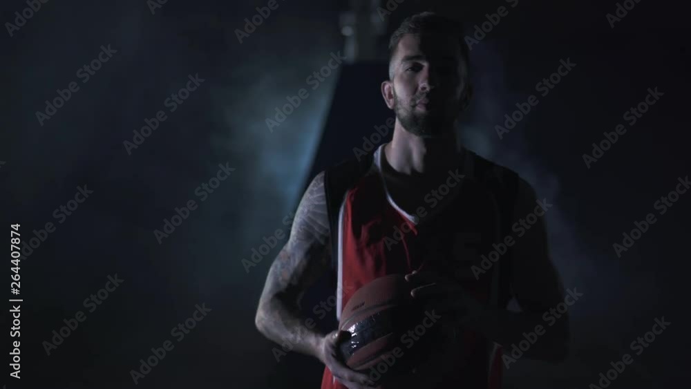 Portrait young handsome brutal unshaved confident basketball player stand with a ball in the clouds of smoke on black background. Portrait young handsome brutal unshaved confident basketball player stand with a ball in the clouds of smoke on black background.