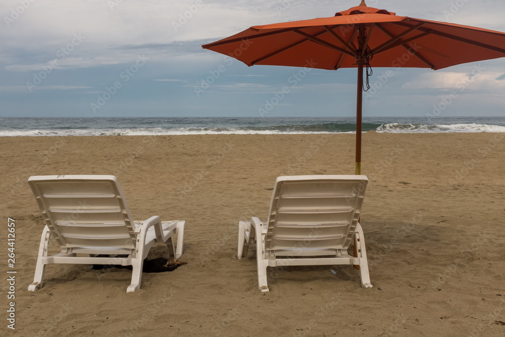 Two empty plastic white chairs under an orange beach umbrella on a deserted sandy beach in Costa Rica
