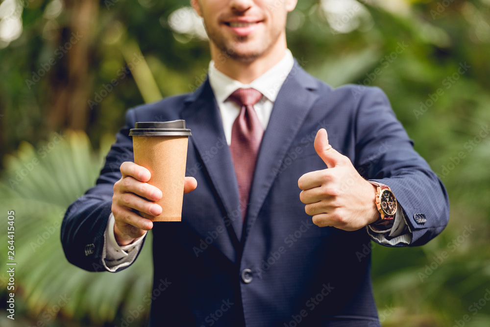 cropped view of smiling businessman in suit and tie holding coffee to go and showing thumb up in orangery cropped view of smiling businessman in suit and tie holding coffee to go and showing thumb up in orangery