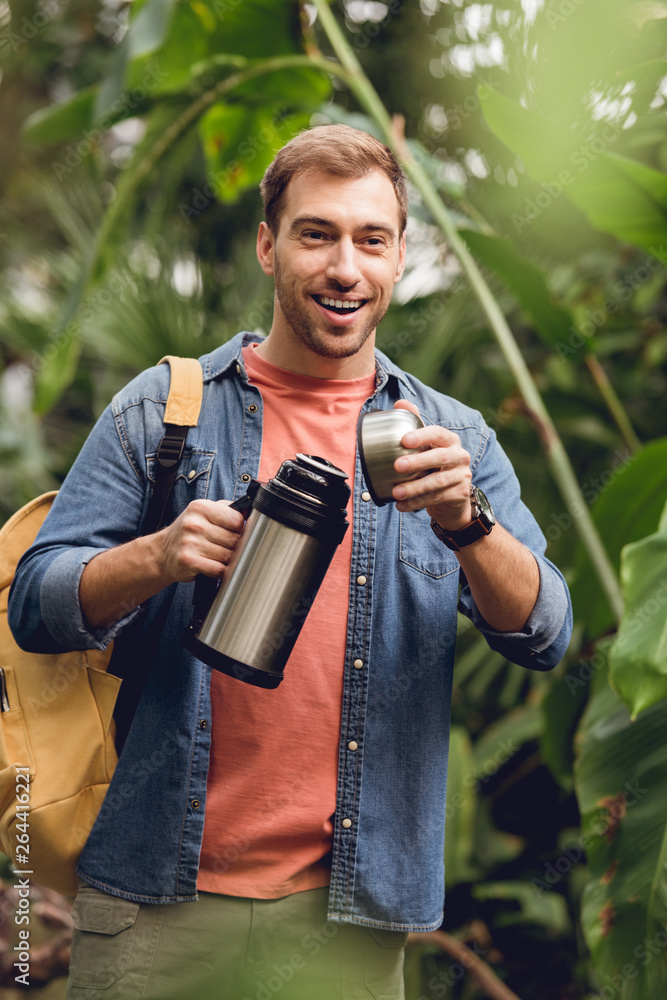 Naklejka premium selective focus of happy traveler with backpack opening thermos in tropical forest