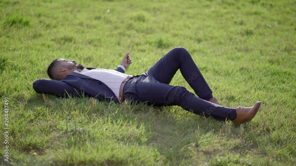 Man in suit lying on green lawn. High angle view of handsome relaxed businessman resting on green grass, full length view, handheld shot. Relaxation concept Man in suit lying on green lawn. High angle view of handsome relaxed businessman resting on green grass, full length view, handheld shot. Relaxation concept