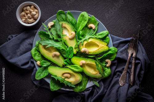 Sliced ​​avocado slices, arugula salads, spinach and cashew nuts on a gray plate and gray linen cloth against a black background. top view
