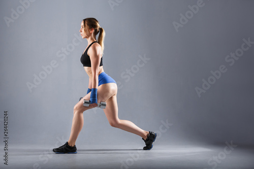 Side view of a beautiful young woman fitness instructor wearing a sports suit - shorts and a top doing an exercise lunges with dumbbells on a gray background
