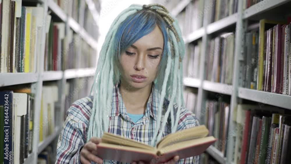 Young cheerful hipster woman with dreadlocks reading book in front of the camera in the library room. Young cheerful hipster woman with dreadlocks reading book in front of the camera in the library room.