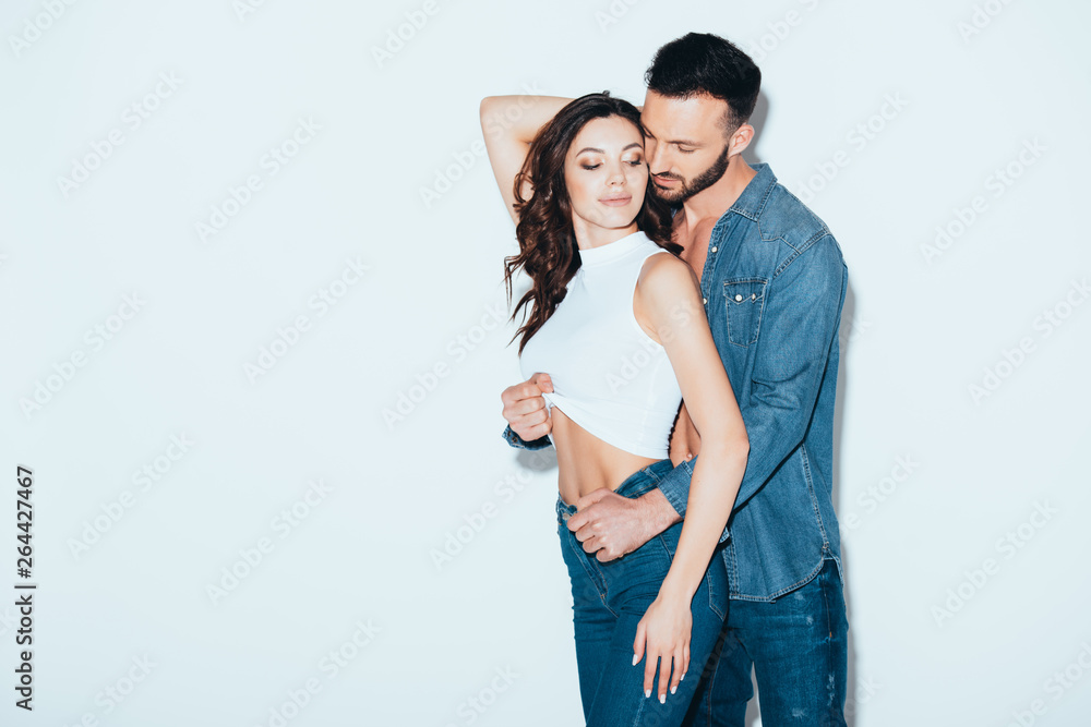 Naklejka premium bearded man in jeans undressing girlfriend on grey