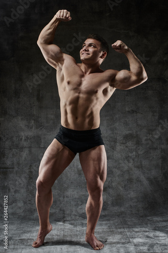 Sexy bodybuilder man posing in full growth on dark background in black shorts. Handsome pumped male body isolated with free space for advertising