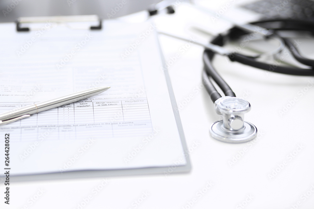 Stethoscope, clipboard with medical form lying on hospital reception desk with laptop computer and busy doctor and patient communicating at the background. Medical tools at doctor working table Stethoscope, clipboard with medical form lying on hospital reception desk with laptop computer and busy doctor and patient communicating at the background. Medical tools at doctor working table