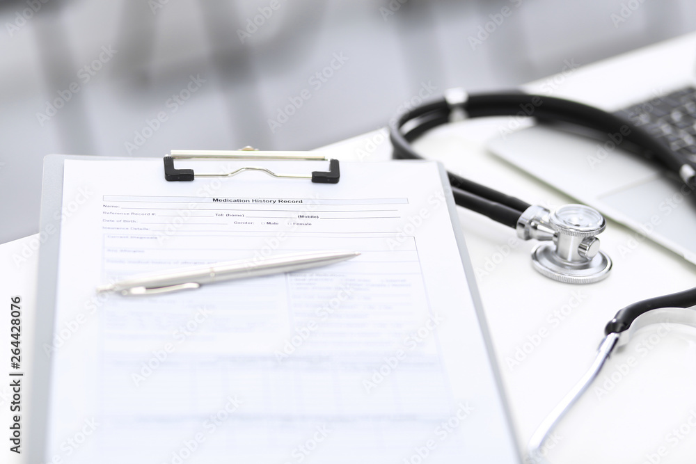 Stethoscope, clipboard with medical form lying on hospital reception desk with laptop computer and busy doctor and patient communicating at the background. Medical tools at doctor working table Stethoscope, clipboard with medical form lying on hospital reception desk with laptop computer and busy doctor and patient communicating at the background. Medical tools at doctor working table