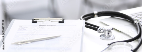 Stethoscope, clipboard with medical form lying on hospital reception desk with laptop computer and busy doctor and patient communicating at the background. Medical tools at doctor working table photo
