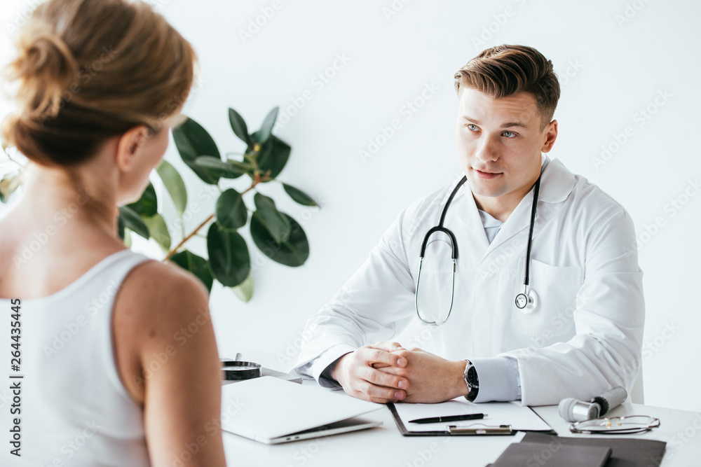 selective focus of doctor in white coat sitting with clenched hands and looking at patient selective focus of doctor in white coat sitting with clenched hands and looking at patient