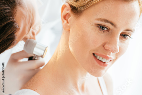 selective focus of attractive woman smiling while dermatologist examining neck with dermatoscope
