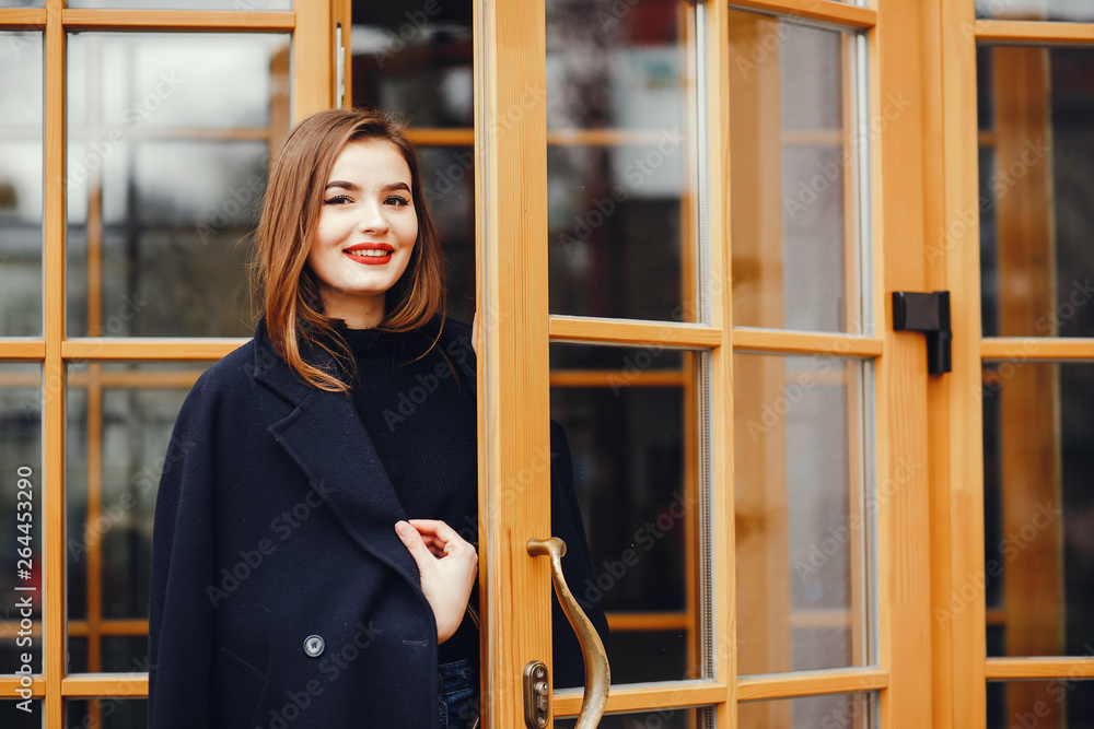 Beautiful girl in a black coat. Cute lady near building. Woman at the door