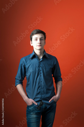 Handsome confident man in blue shirt with rolled sleves holding hands in pokets and looking at camera on orange background