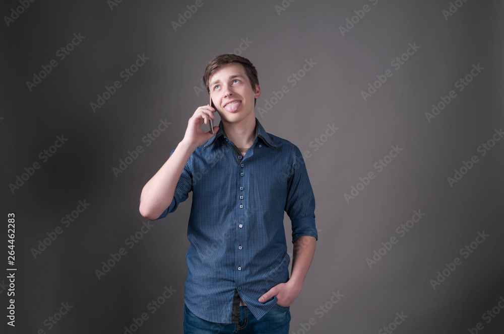 confused handsome young man in blue shirt with sticking out tongue talking on smartphone on grey background with copy space