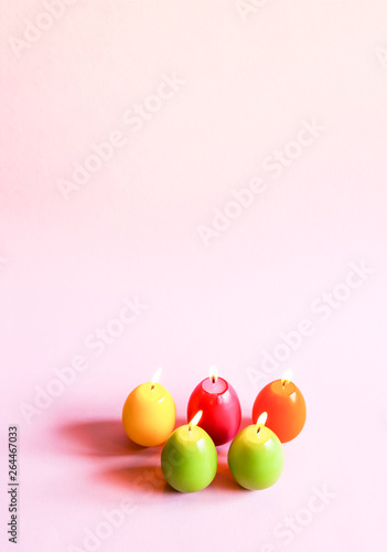 Traditional Easter decor. Group of bright burning paraffin candles in the shape of colorful eggs on pink background.