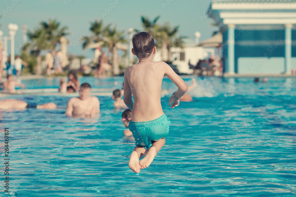 Caucasian boy having fun making fantastic jump into swimming pool at resort. He has pose of sitting man. Back view.