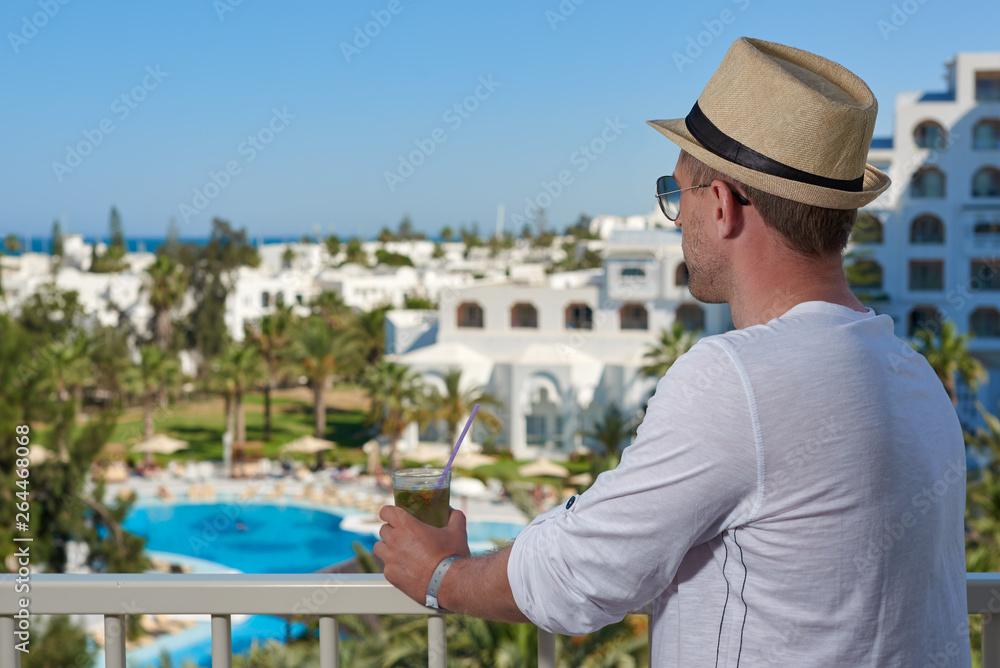 European Man in sunhat with glass of long drink cocktail. He is standing on the hotel’s balcony and enjoying picturesque view.