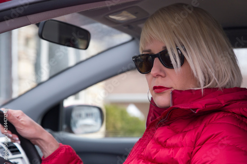 Low angle view of pretty serious platinum blonde young woman dressed in red and wearing sunglasses framed by an open car window