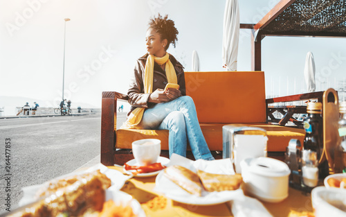 A wide-angle view of a young beautiful African-American female having breakfast in an outdoor cafe; a dazzling Brazilian girl on an orange bench in a street bar having brunch with food in front of her