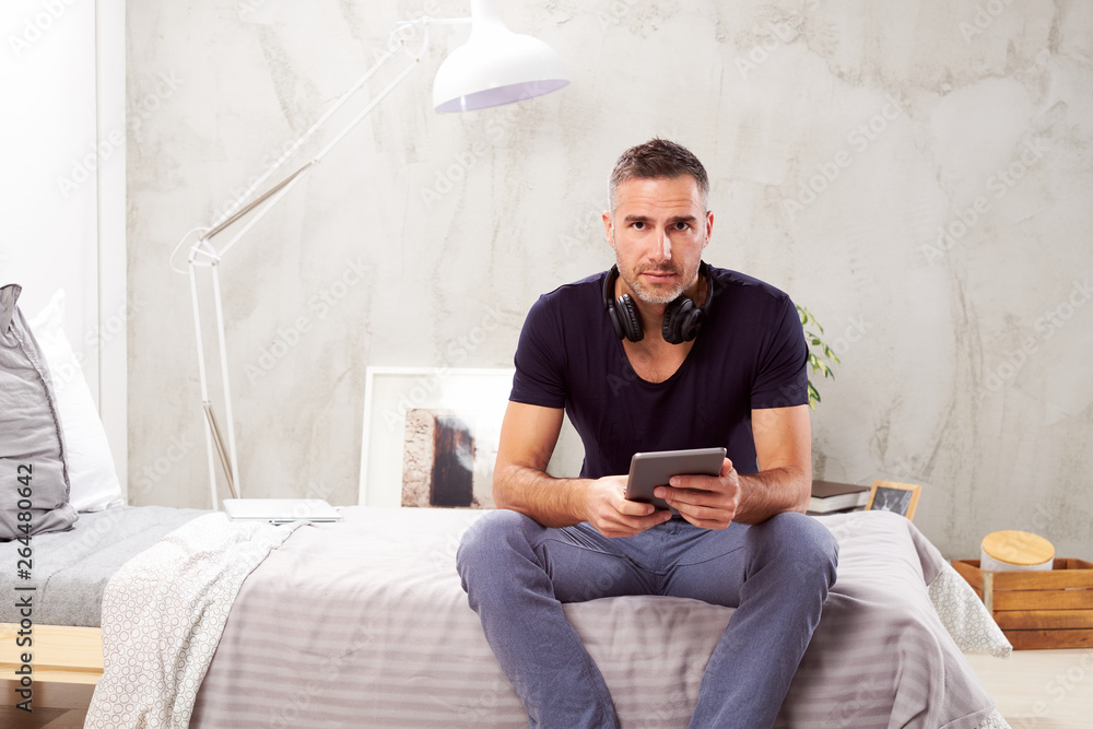 Caucasian man in forties sitting on bed in bedroom and using tablet.