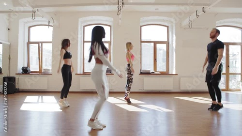 Tracking right shot of three young women doing jumping jacks to the music during group fitness training. Male instructor showing exercise photo