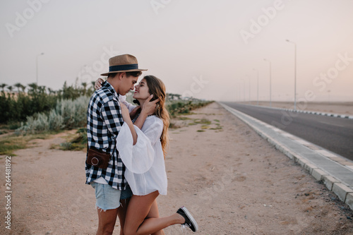 Graceful pretty girl in white tunic standing on one leg and gently embracing her boyfriend near the road. Young man in denim shorts and trendy hat looking at his girlfriend's eyes on outdoor date © Look!