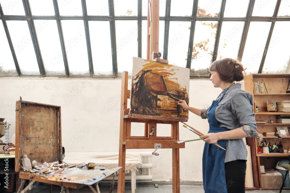 Woman artist paints a picture on canvas. Bright art studio with a large window. Easels and canvases. Woman artist paints a picture on canvas. Bright art studio with a large window. Easels and canvases.