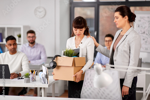 business, firing and job loss concept - colleague seeing off sad fired female employee leaving office with box of her personal stuff