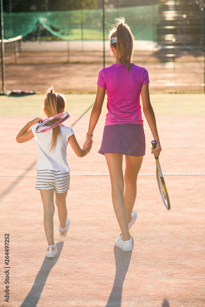 Female tennis trainer holds her little student to the hands and they goes together after training at outdoor tennis court at sunset.